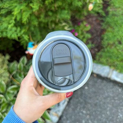 A hand holding a Ceres Chill Chiller with a Coffee Slider lid attached with green plants in the background. 